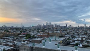 The Philly skyline at a cloudy dusk, tiny near the horizon, over the teeming roofs of South Philadelphia.