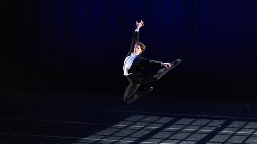 Hollis in a leotard fashioned like a tuxedo leaps in the air, vignetted light panels onto the floor