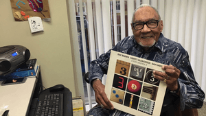 Perkins, an elderly Black man wearing a patterned button-down, sits at a desk smiling and holding a vinyl jazz album.