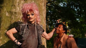Two characters, one in drag, and another looking on at them, eager and smiling, stand outdoors by a large tree