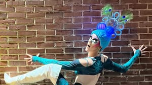 Mayer poses against a brick wall, one leg lifted, in blue peacock-inspired drag and thigh-high white stiletto boots.