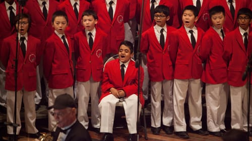 Close-up on about 15 members of the Boys Choir, wearing white pants and red blazers, singing together.