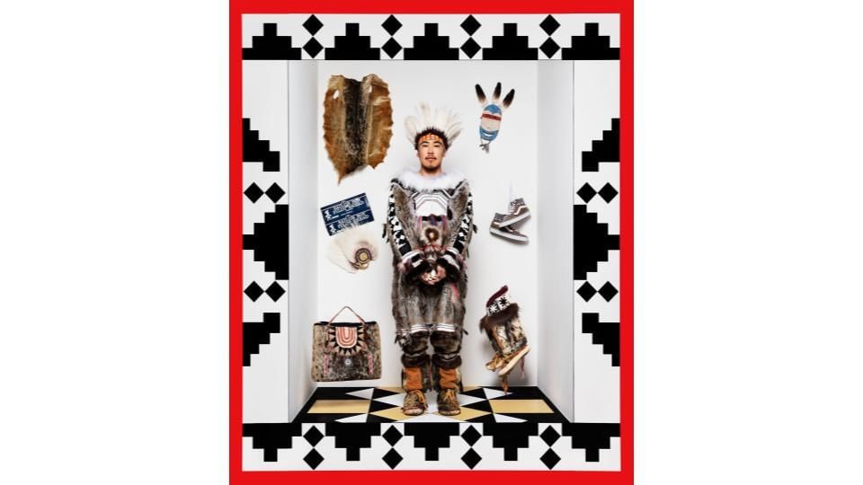 An Indigenous man stands in a life-size frame with bold black geometric designs. He wears an ornate traditional fur outfit