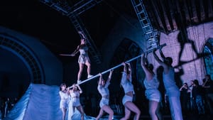 A trapeze artist walks along a pole held up by 6 other dancers in a room with a dim, violet-purple tinted light