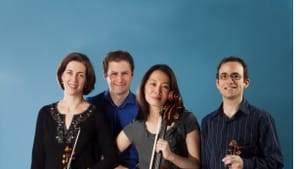 The quartet members, grouped together in dark clothes, smiling and holding their instruments, against a blue background.