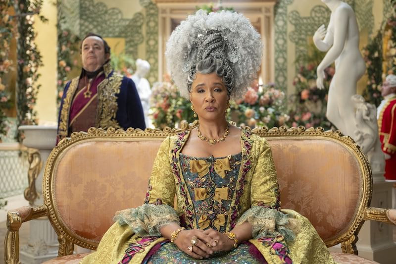 Sachs, an older white man, attends a lavishly dressed Rosheuvala, a Guyanese English woman wearing a towering royal gray wig.