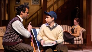 Scene from the play. Eugene, wearing a cream knit sweater, argyle socks, & baseball hat, looks at his brother, who’s speaking