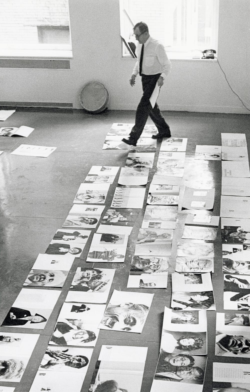 Informal black & white daytime photo of Brodovitch walking across dozens of photos laid flat on the office floor.