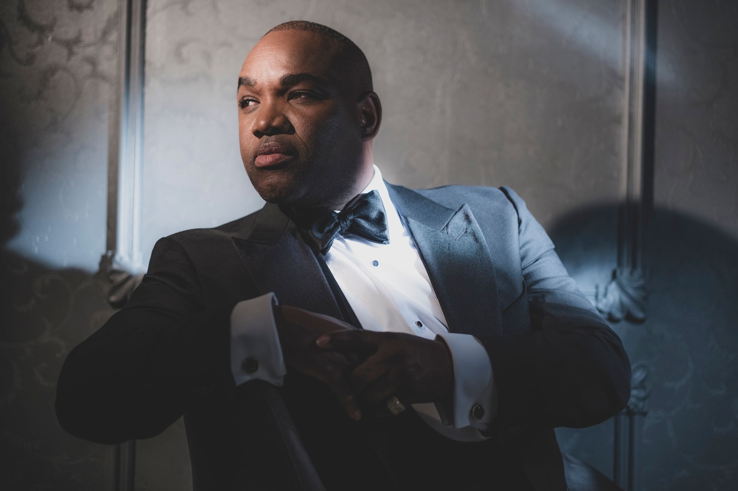 Brownlee, a Black man in a dramatic shadow, strikes a contemplative pose in an opulent blue-gray tuxedo.