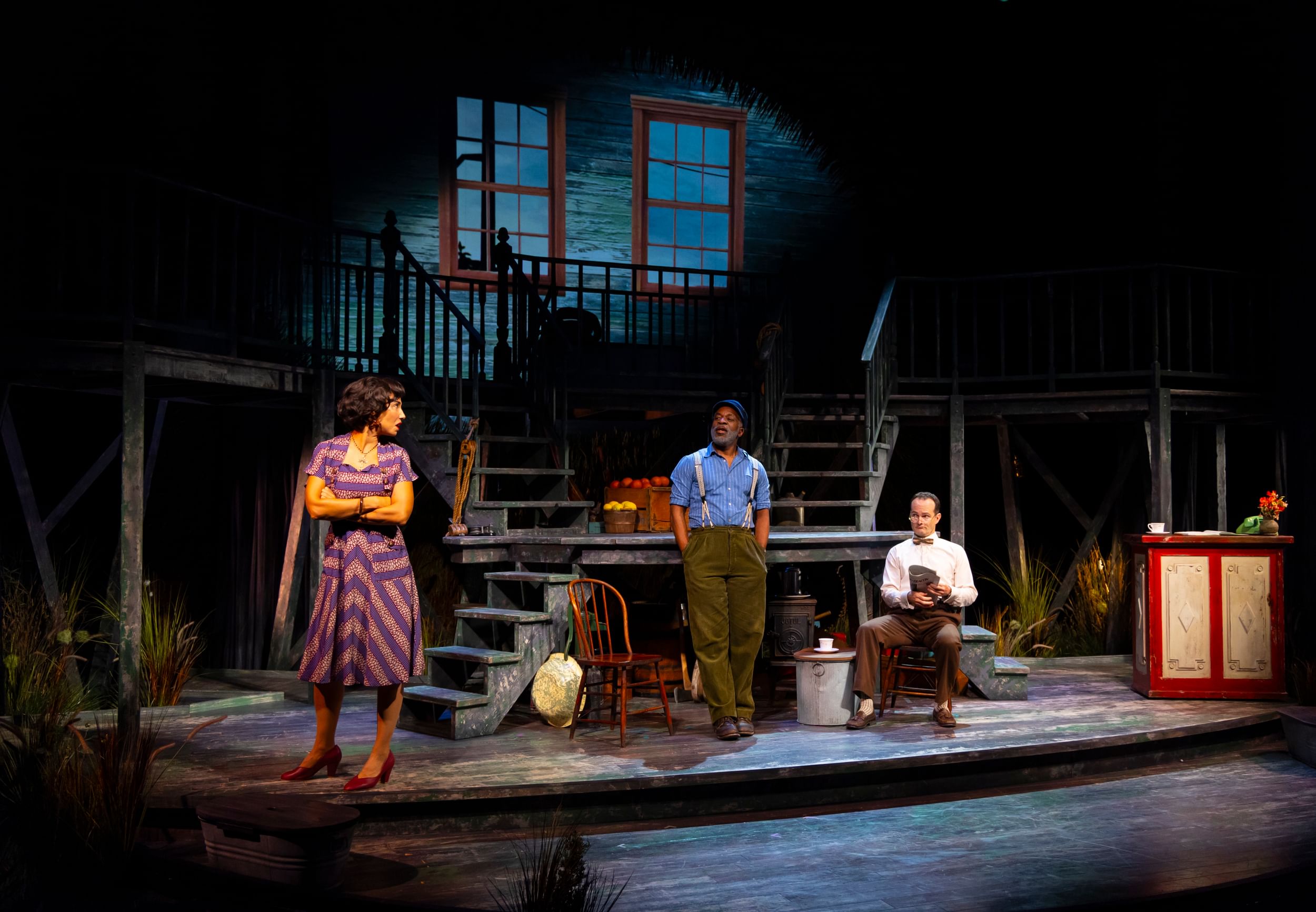 Scene from the play showing richly textured 3-level set with chairs, fruit crates, stairs, bannisters & glowing blue windows