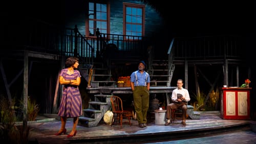 Scene from the play showing richly textured 3-level set with chairs, fruit crates, stairs, bannisters & glowing blue windows