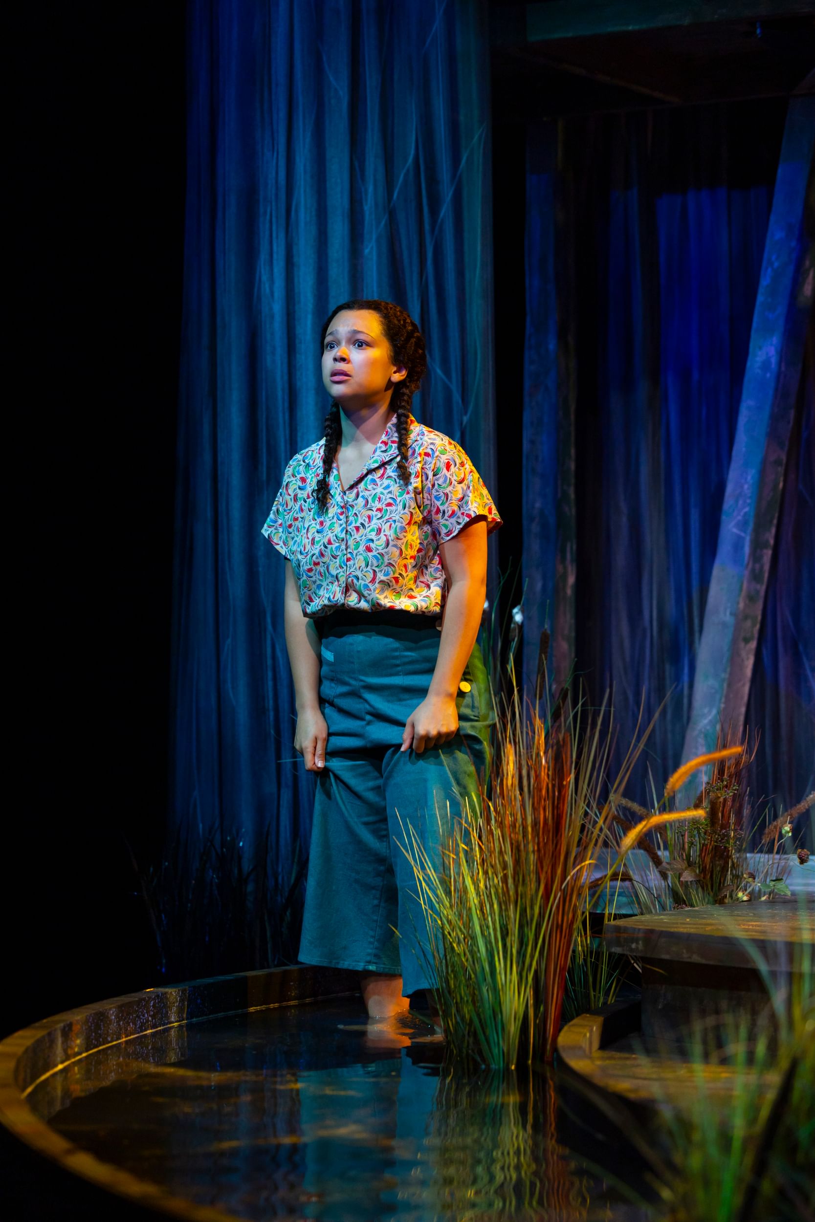 Tyson, in colorful button-down & wide blue capri pants, wades in a reedy waterway created onstage.