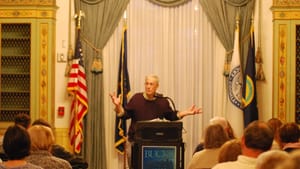 Christopher Bursk, a white man w/ white hair, speaks at a college lectern with arms outstretched, to a crowd seated in front