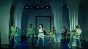 Ensemble of 5 women strike the same pose, wearing toga-like white shirts, among white fabric stretched like classical columns