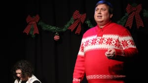 A man in a bright red Christmas sweater on stage speaks towards audience, an actor to his left is sitting, crying