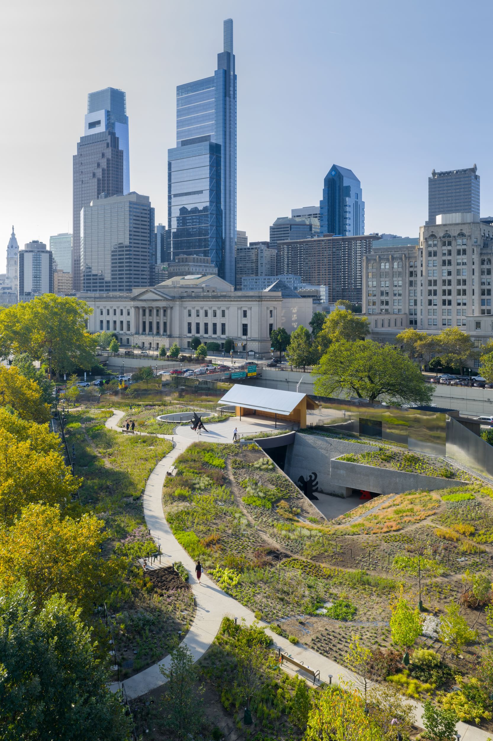 Aerial view of the gardens showing curving paths, green gardens, and trees, with the Philly skyline rising close beyond.