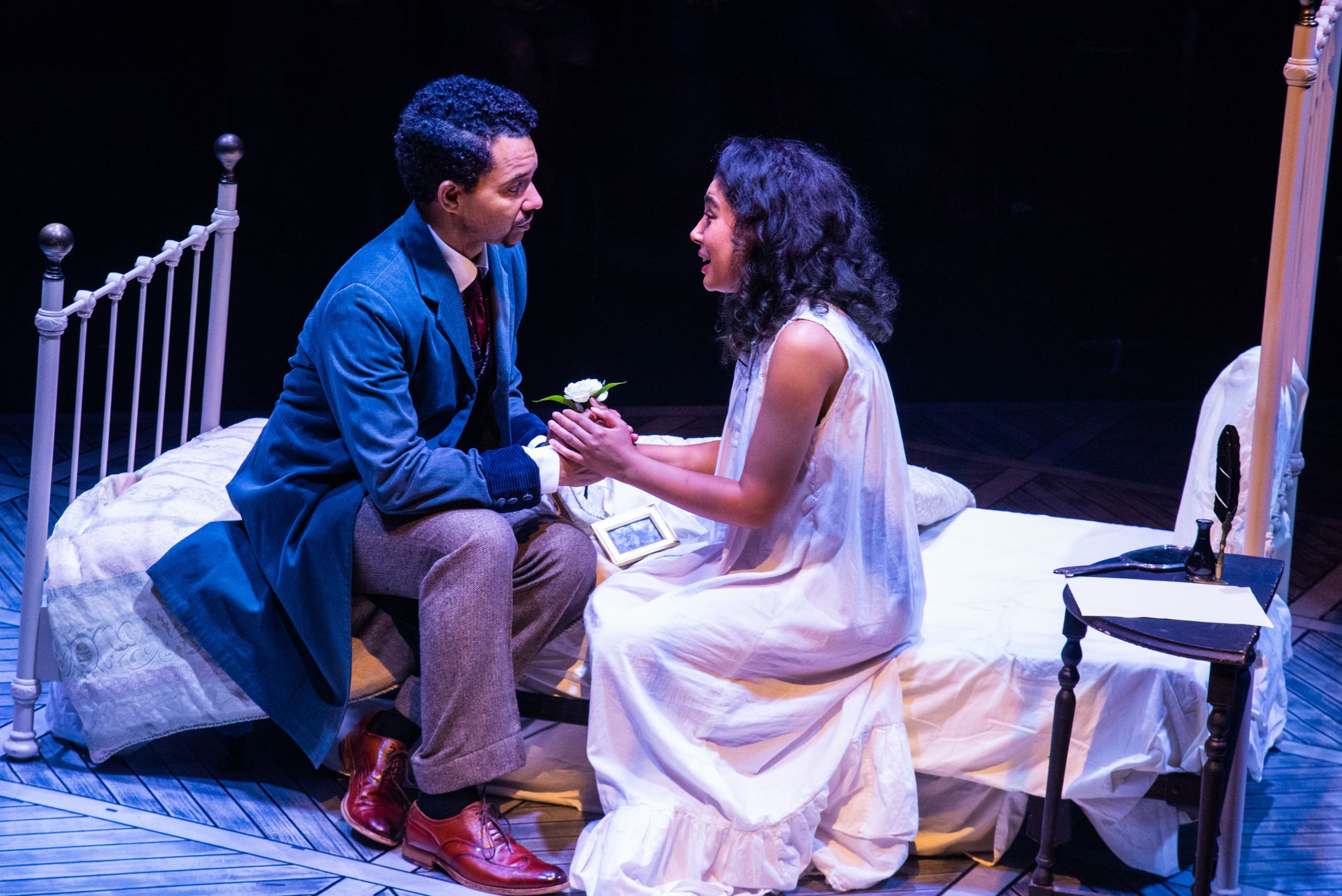 Scene from the play. Richardson & Wyatt, two Black actors, face each other tenderly with hands joined, sitting on a small bed