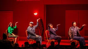 In contemporary clothes in different styles, the singers sit sideways in a row on chairs, gesturing up with their left hands.