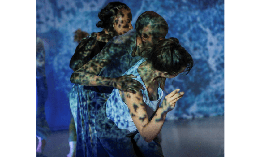 Under a lace-like blue lighting effect, three dancers bend gently forward while pressed against each other, front to back.