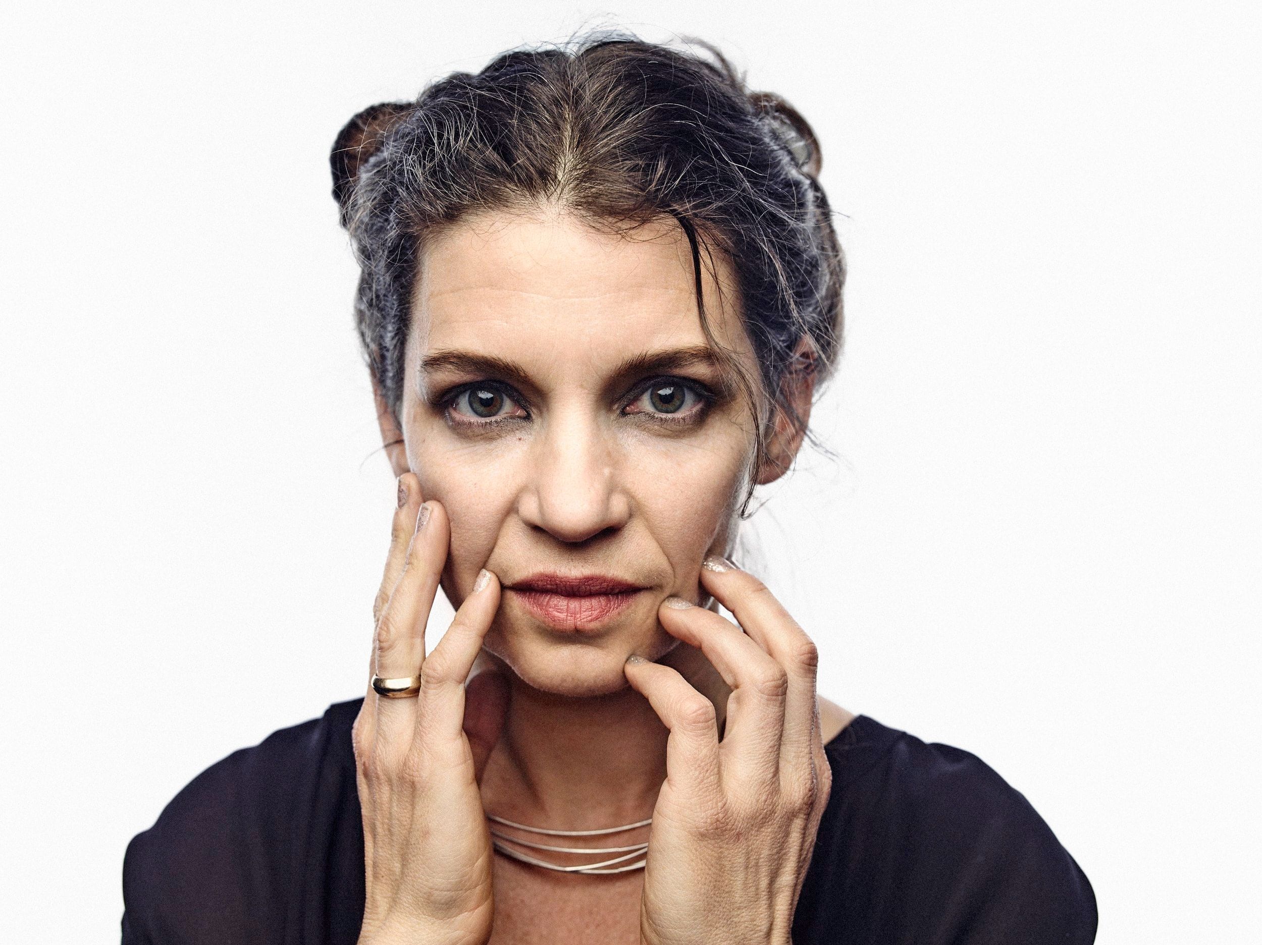 Close up photo of Kihlstedt, her hands at her face as she looks deeply towards camera, in front of all white background