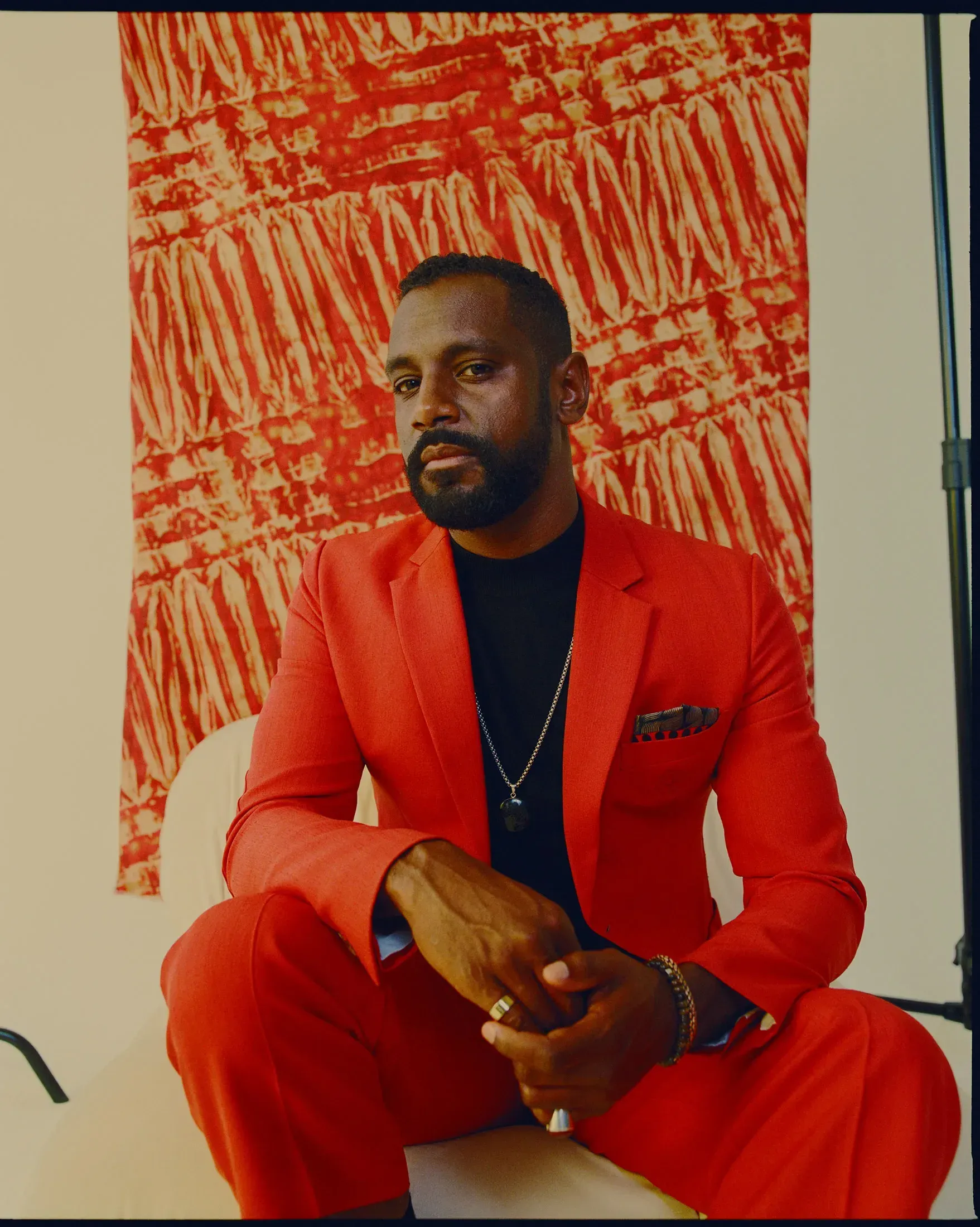 Simon, a Black man, in a red-orange suit with black shirt and necklace, poses for portrait in front of a red-orange tapestry