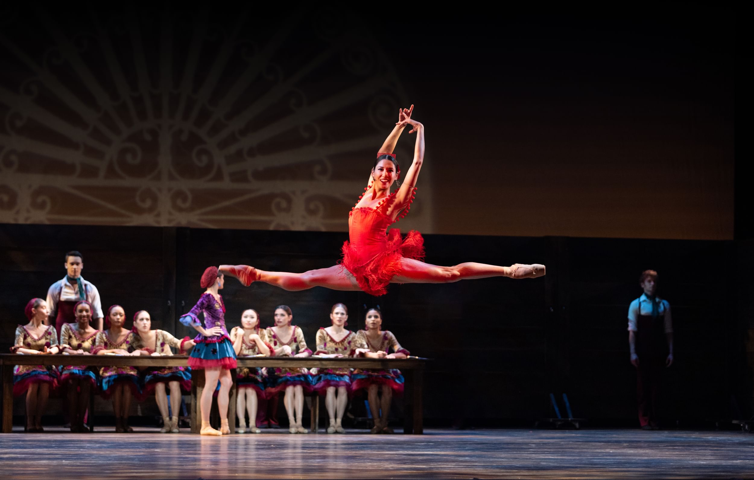 A dancer leaps into the air doing a full split, wearing all red, other artists on stage behind her