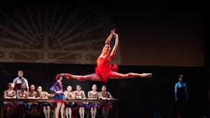 A dancer leaps into the air doing a full split, wearing all red, other artists on stage behind her