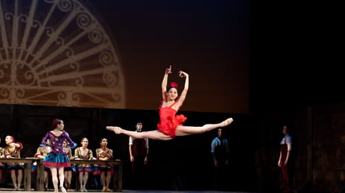 With a giant fan project on the back wall, a dancer in a red tutu and leotard leaps high with her legs and arms extended.