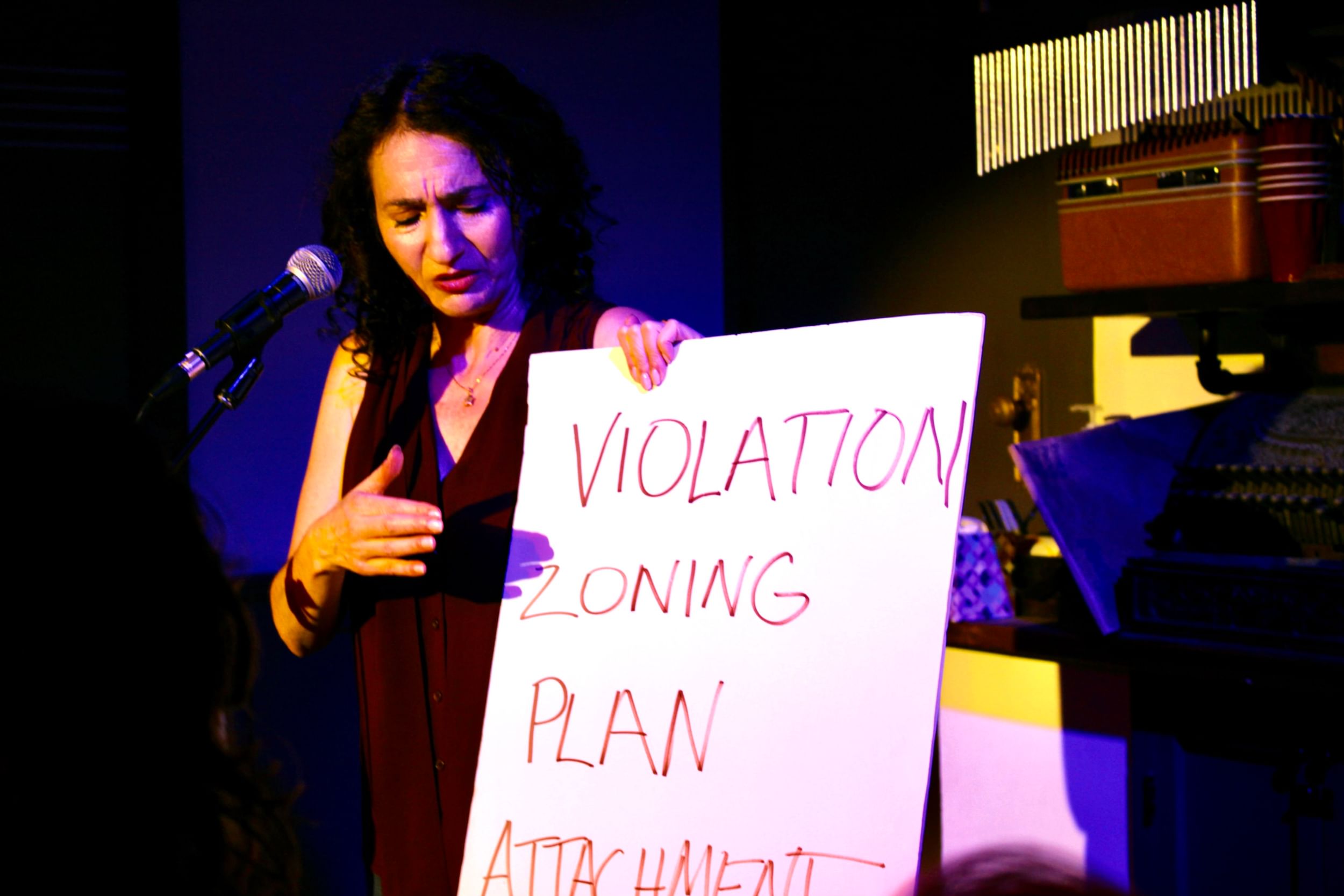 A woman on stage by a microphone holds a white board with the words “Violation Zoning Plan Attachment” written large, in caps