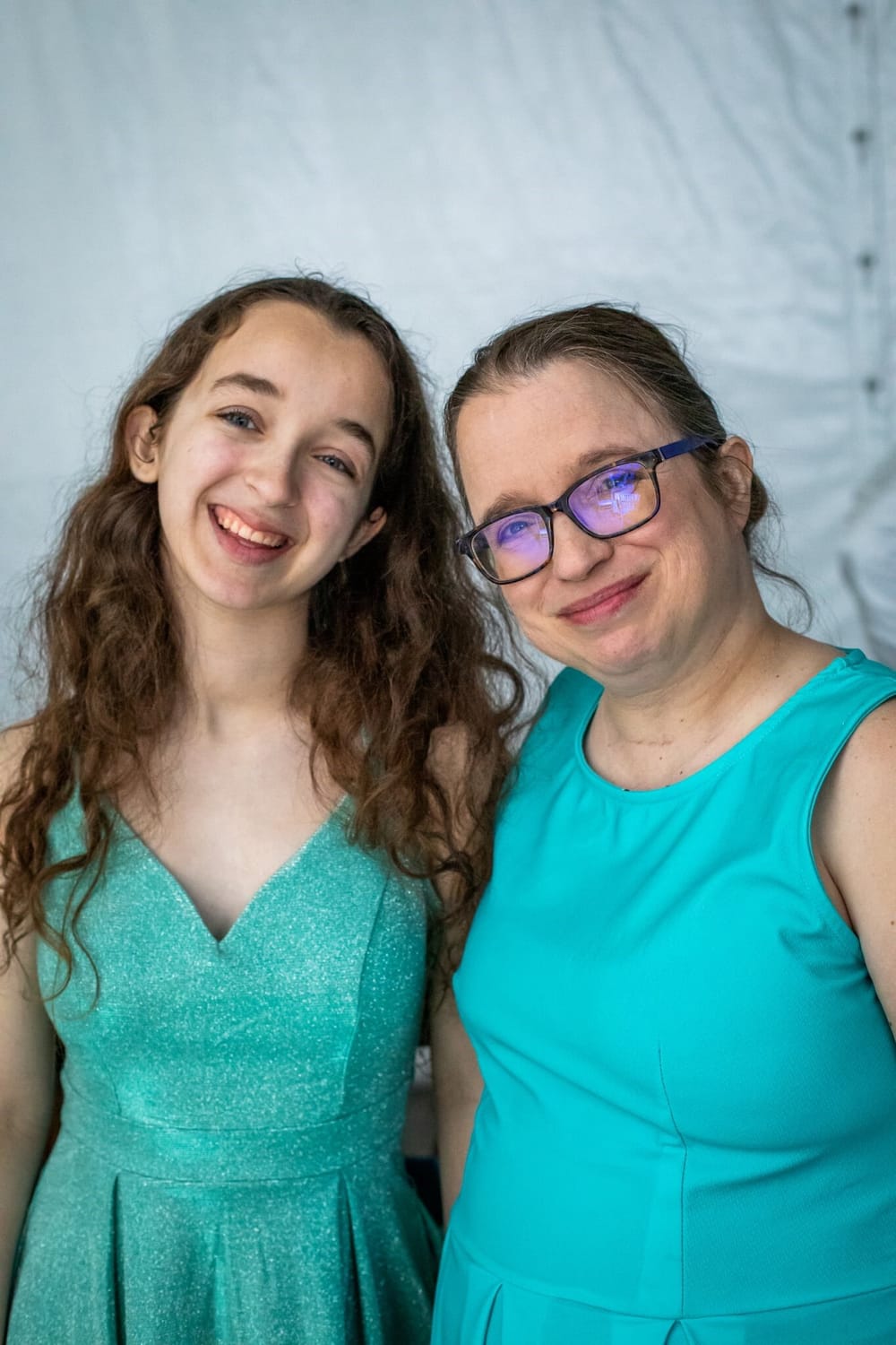 Carrie, a white woman in her 40s with glasses, stands smiling with her teen daughter. Both wear turquoise dresses.