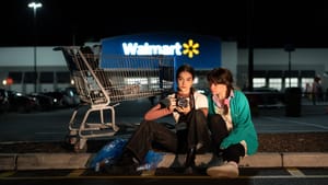 A nighttime photo of characters Tess and Rach in a Walmart parking lot, next to a shopping cart. Tess holds a camera.