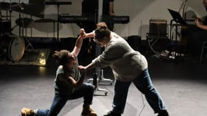 In front of a small band, the two people seem to wrestle, their hands joined pushing each other, Painter down on one knee.