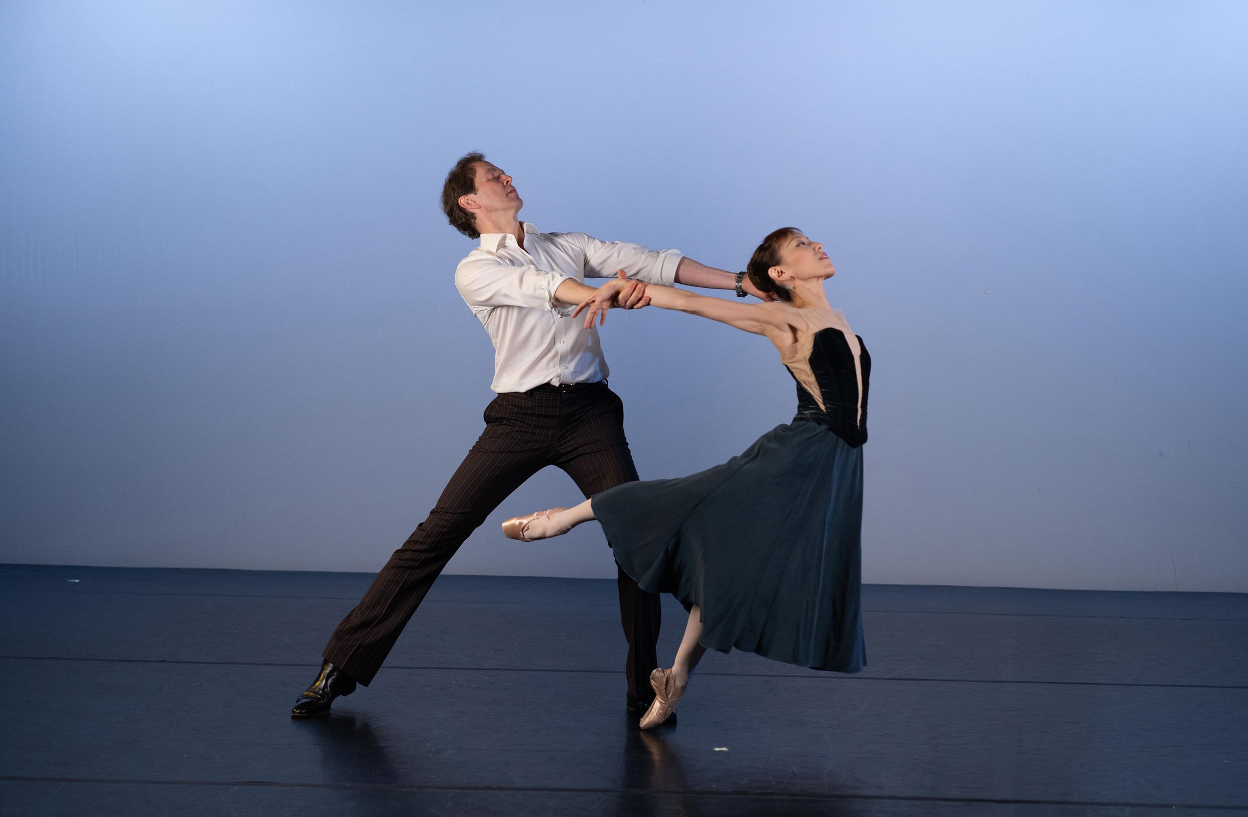 Two dancers in a rehearsal space, one dancer holds the other by the arms as she extends out, her leg kicked back