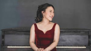 Hu in a red velvet suspender dress sits in front of a piano, one hand over other, head turned left, slight smile in pose
