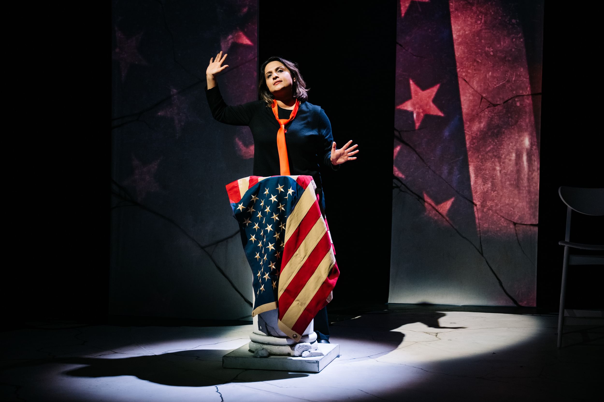 Shetty, an Indian woman in black top and loose orange tie, gestures with her palms out over a US-flag draped lectern.