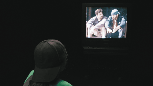 A child with a backward baseball cap in a dark room watches Chasing Amy on a square 90s TV.
