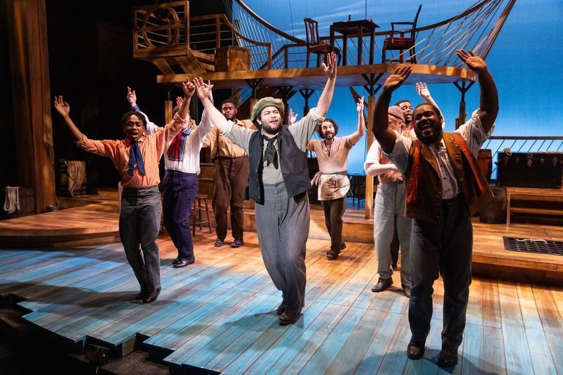 Multiracial group of 9 people in 19th-century sailor clothes sing together on a set like the deck of a ship, arms raised