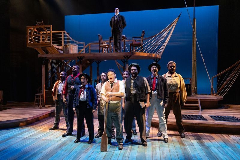 Nine people dressed as sailors pose in a tight group, singing, on a set like the deck of a ship with a deep blue backdrop.