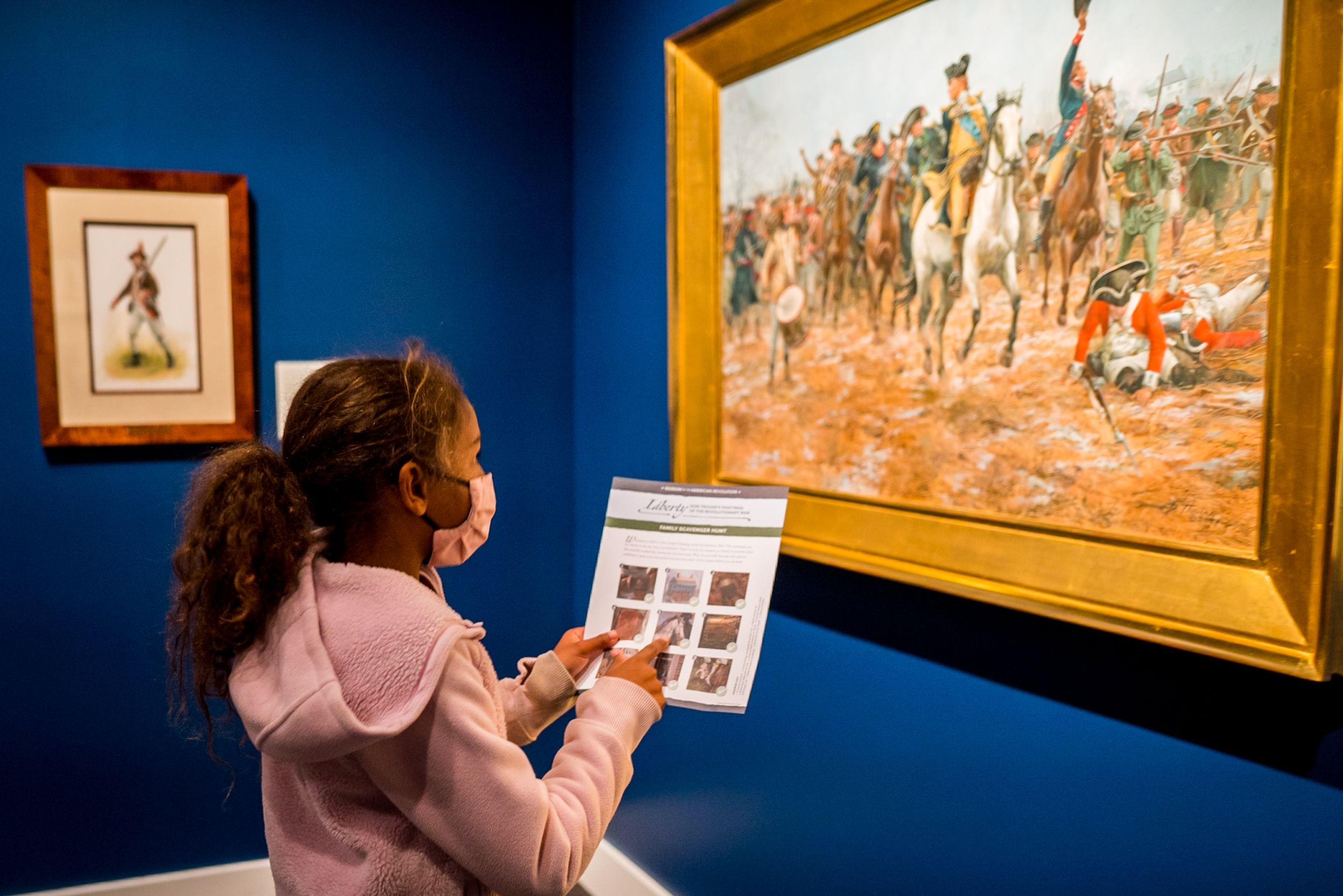 A young girl looks on at a framed illustration on the wall. She's holding a guide to go along with the painting