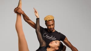 Two Black dancers in pas de deux, wearing all black with shirts that have Chocolate Ballerina Company printed on them