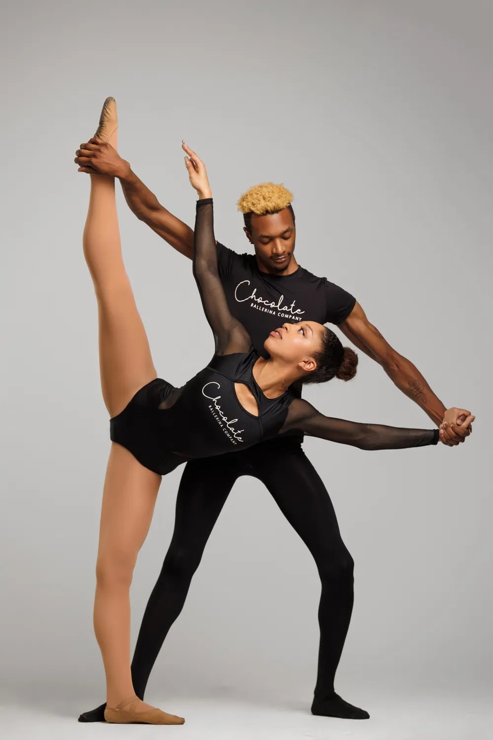 Two Black dancers in pas de deux, wearing all black with shirts that have Chocolate Ballerina Company printed on them