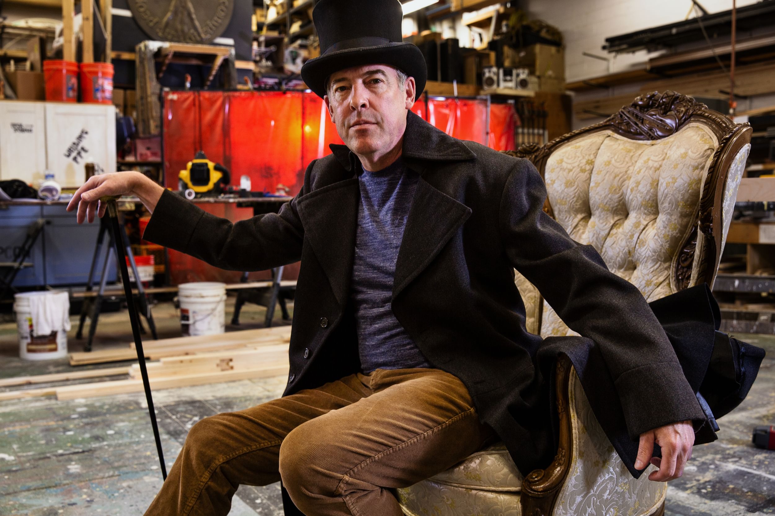 Merrill in a peacoat, top hat, and with a cane sits in a cushioned chair, a set being built behind him.