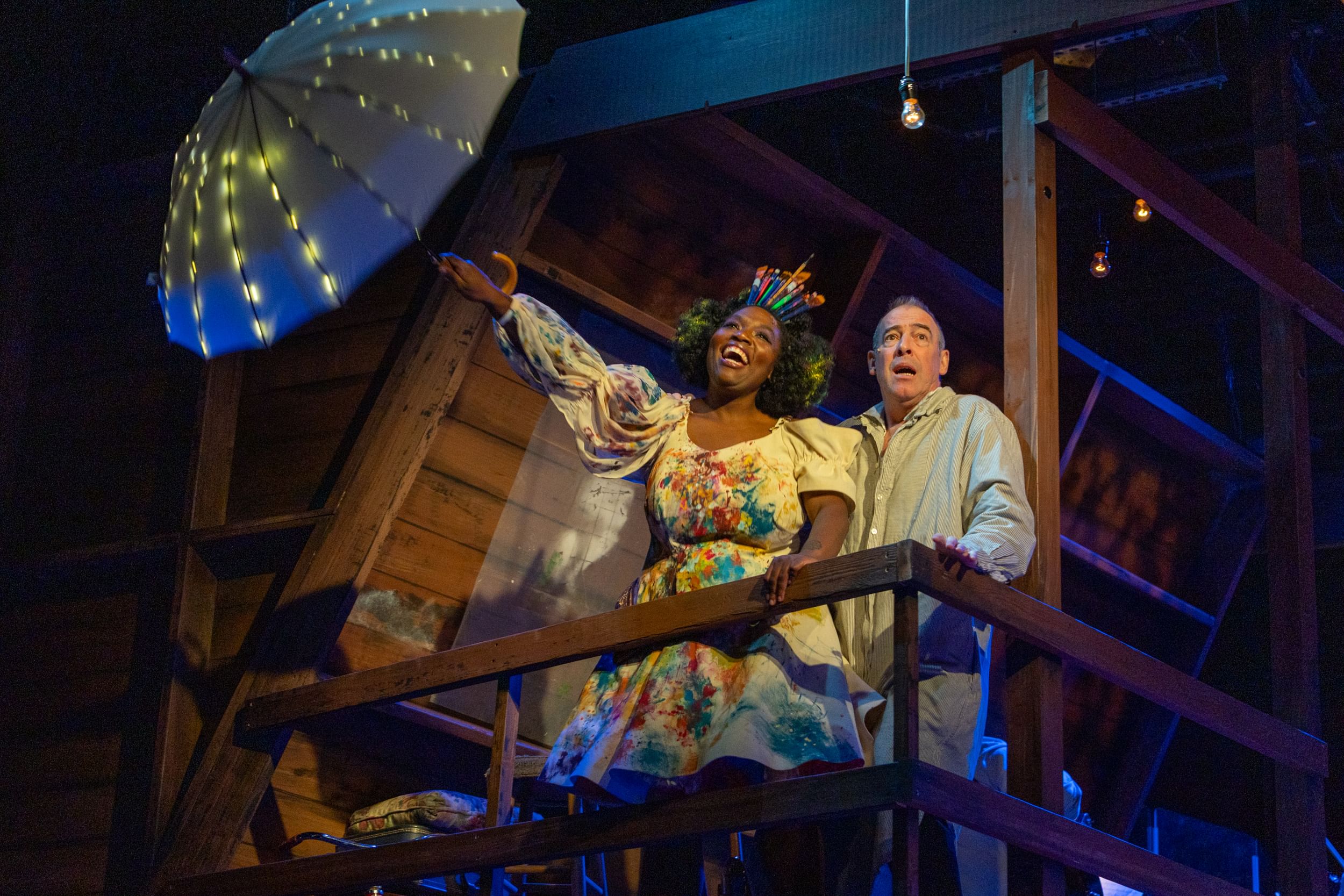 Seen from below in a wooden attic, Williams leans joyfully out holding a lighted umbrella while Peakes looks frightened.