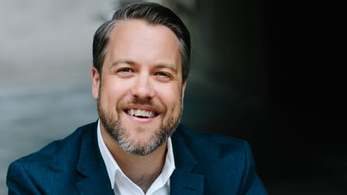 A photo of Christopher Gruits, a white man in a blue suit. He sits with his elbows on knees with his hands joined, smiling.