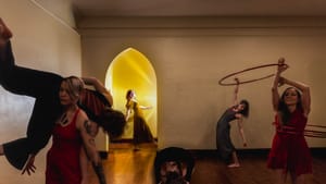 In a gold-lit room with an arched doorway, six people pose gracefully, some with hula hoops.