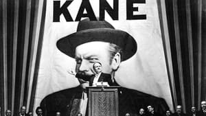 B&W film still. A man in a suit at a podium, other men in suits look on behind him, a large poster with a man reads KANE.