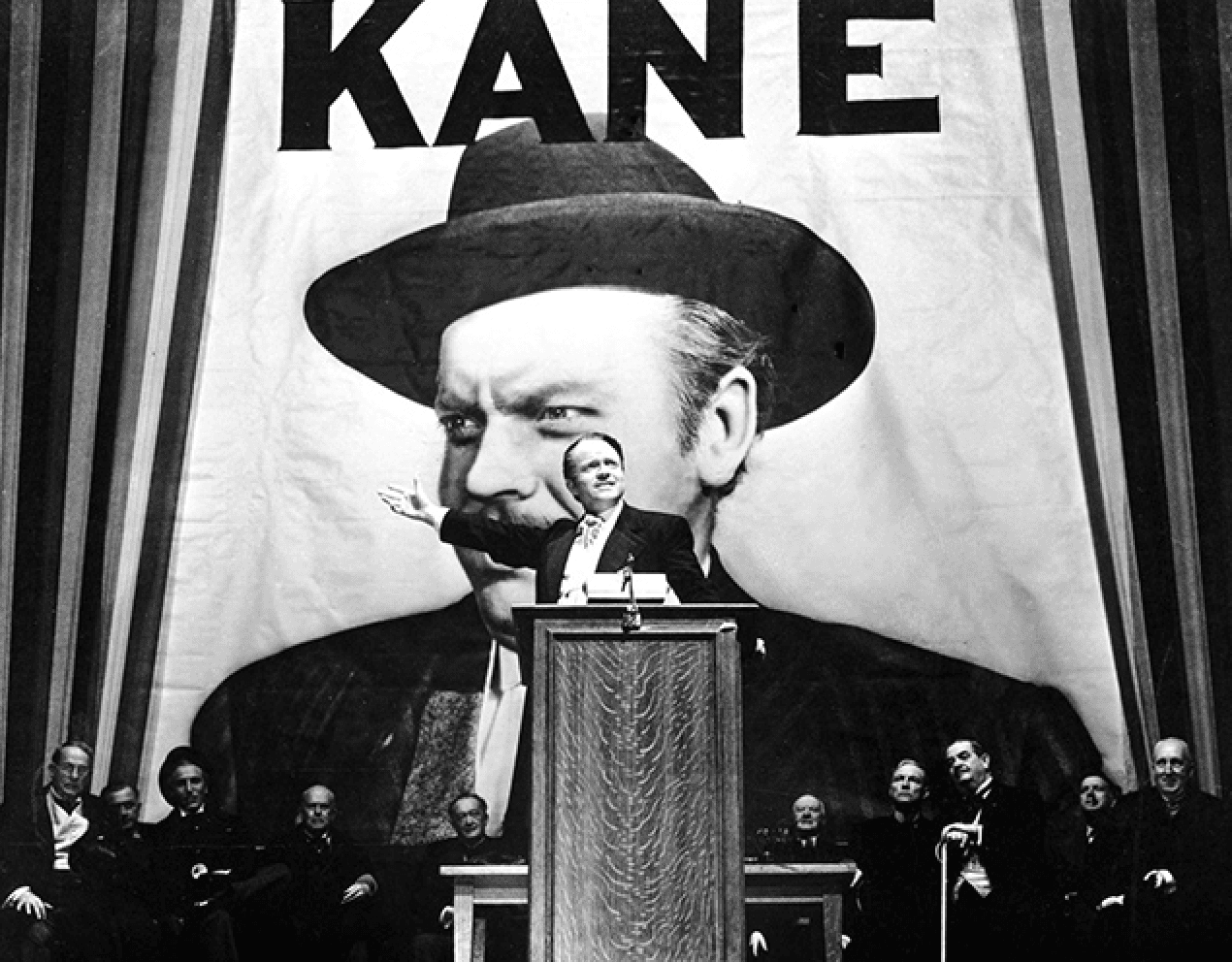 B&W film still of a man in a suit at a podium gestures out to the audience off camera, several men in suits behind him.