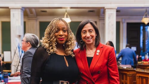 Gauthier, wearing a black dress, stands smiling with Landau, wearing a red blazer, in City Council chambers.