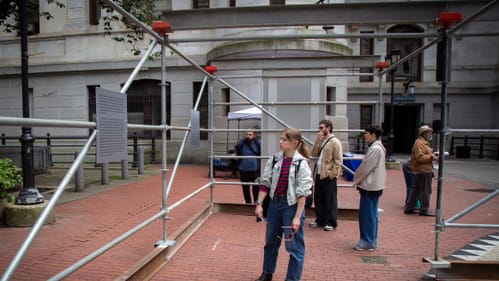 Four people in casual outdoor clothes look at large metal plaques on a metal scaffold in the City Hall Courtyard.