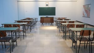 View from the back of a small bare classroom with rows of empty desks facing a blank green chalkboard.